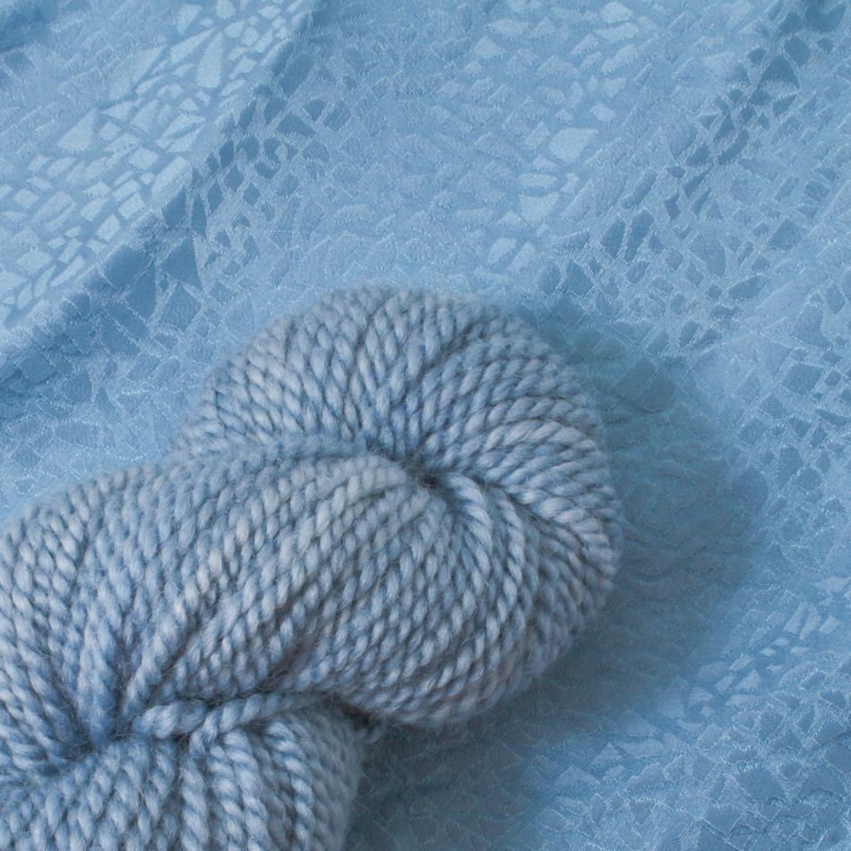 A skein of thick, soft blue yarn is placed on a textured blue fabric surface. The fabric, dyed using Earthues Indigo by Earthues, features an intricate, lace-like pattern that contrasts with the yarn's smooth and uniform appearance. Both the yarn and fabric share a similar light blue hue.