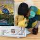 The Felting Studio's Needle Felting Tapestry Kit, placed on a wooden surface, includes comprehensive needle felting instructions and an array of wool in black, green, blue, yellow, and brown. The kit enables you to create a beautiful felted image of a heron standing on rocks near reeds with a golden sky background.