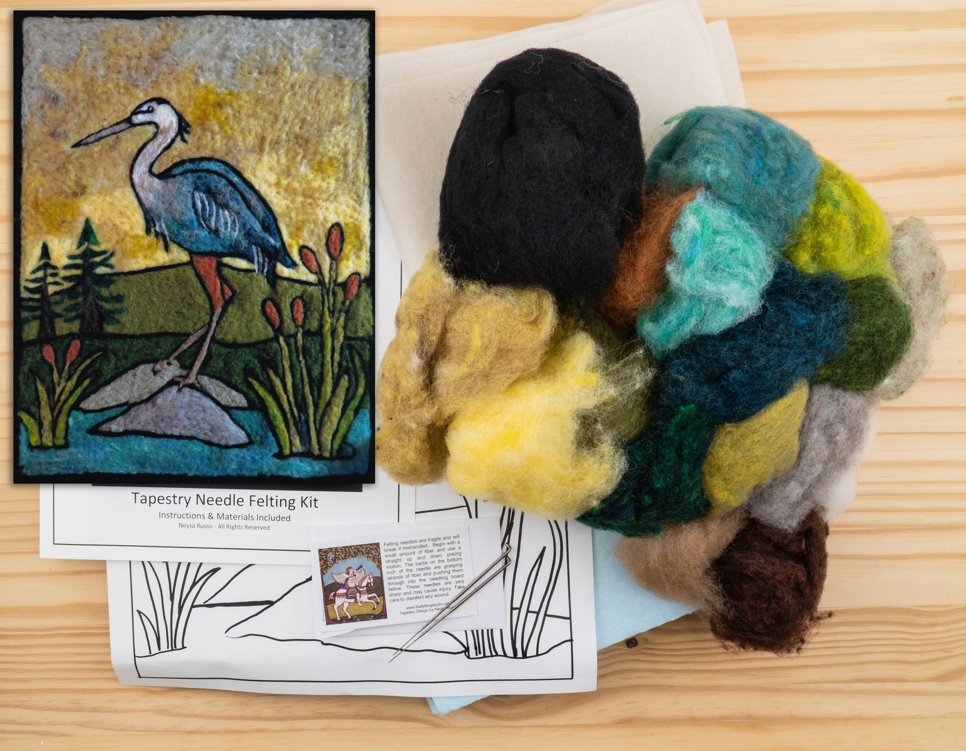 The Felting Studio's Needle Felting Tapestry Kit, placed on a wooden surface, includes comprehensive needle felting instructions and an array of wool in black, green, blue, yellow, and brown. The kit enables you to create a beautiful felted image of a heron standing on rocks near reeds with a golden sky background.