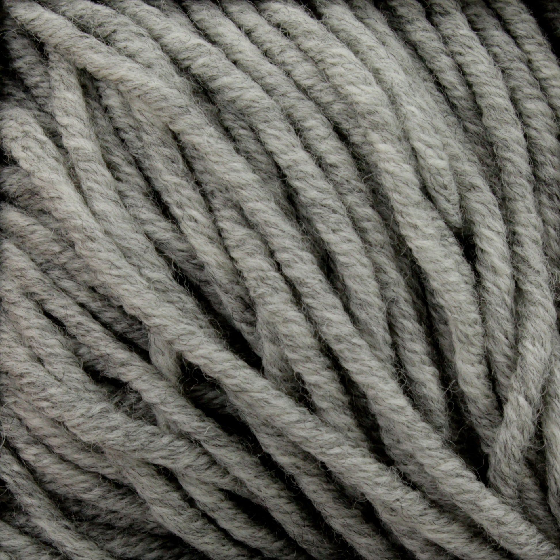 A close-up image of Plymouth Select Worsted Merino Superwash yarn by Plymouth Yarn Co. showcases its thick, twisted grey strands. The texture appears soft and woolly, with the merino fibers tightly wound together. The color gradient ranges from light to dark grey, creating a subtle variegated pattern that highlights excellent stitch definition.