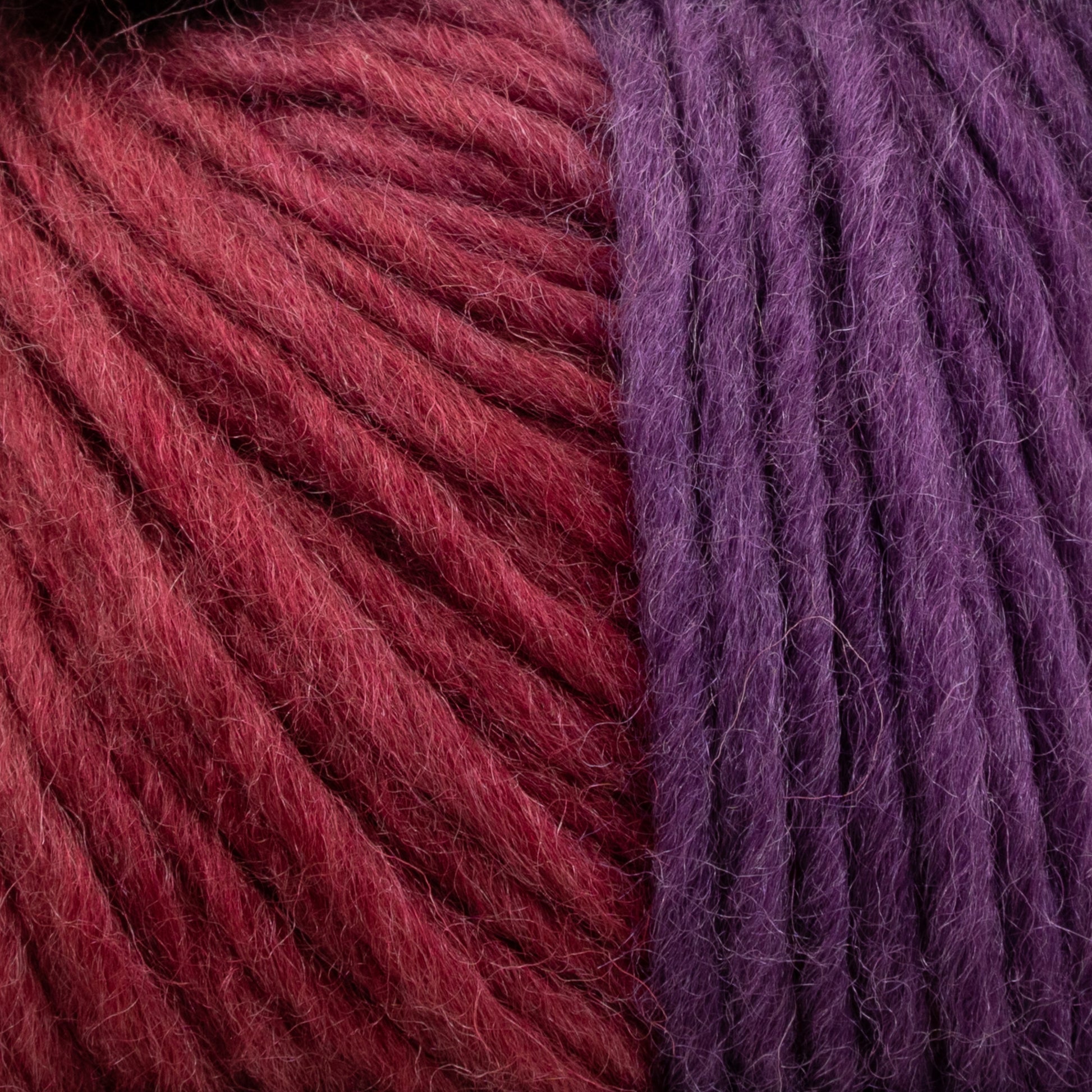 Close-up view of two skeins of Louisa Harding Amitola Grande yarn side by side. The yarn on the left is a deep red color, while the yarn on the right is a rich purple. This worsted weight yarn from Knitting Fever / Euro Yarns has a soft texture, and the fibers appear slightly fuzzy.