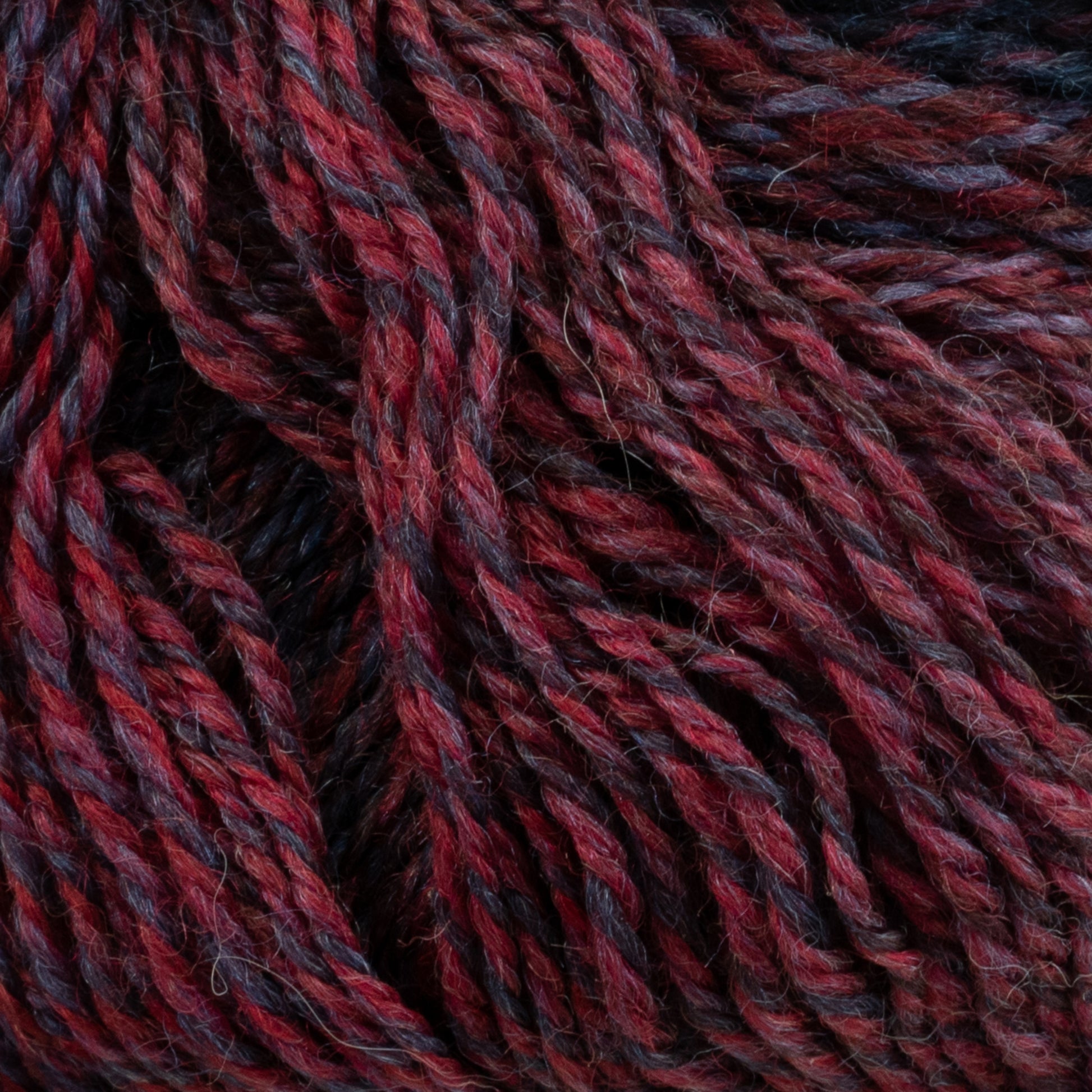 Close-up image of a bundle of intertwined Knitting Fever Painted Desert Yarn by Euro Yarns in shades of red, maroon, and dark gray. The texture is visible, showing the tightly twisted superfine weight fibers and the rich, variegated colors with long color changes, creating a visually appealing pattern perfect for lightweight accessories.