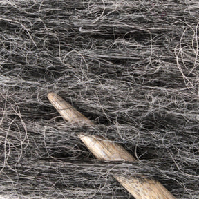 Close-up image of a crochet hook amidst strands of Berroco, Inc. Plötulopi Unspun Icelandic Wool Yarn in gray and black. The hook is partially visible, intertwined with the textured fibers, showcasing the intricate nature of this Icelandic wool.
