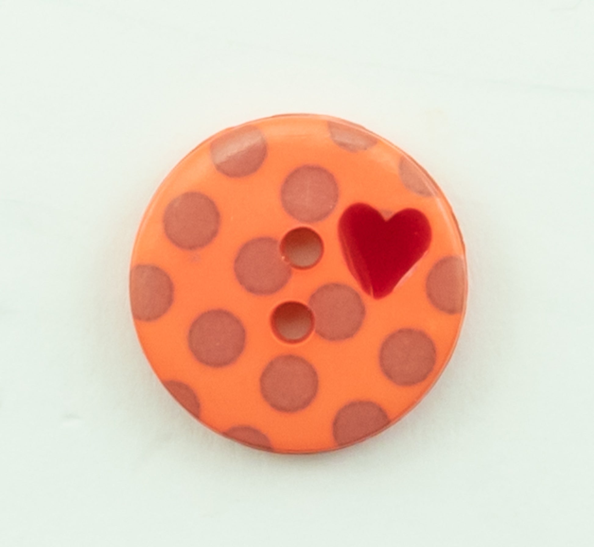 The Dots Button by Buttons Etc. is an orange plastic button with a 5/8" diameter, featuring brown polka dots and two small holes in the center. It includes a red heart design near the upper right side and is set against a light background.