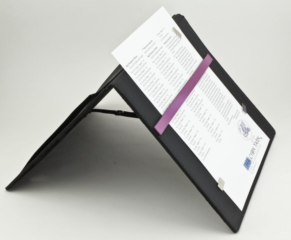 The Magnetic Pattern Holder by Knitter's Pride from Accessories Unlimited, in a sleek black design, stands propped up in a tent-like position, showcasing a white document secured with magnetic clips and a purple band. This efficient fold-up tool ensures that the document, adorned with text and logos, remains intact. The background is plain and light-colored.