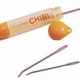 The image shows a Bryson Distributing, Inc. Chibi Bent Tapestry 3 Needle Set with its cap removed. The holder is cylindrical with an orange cap and end. Two easy-to-thread tapestry needles lie next to the holder, showcasing its storage capability and practicality for any sewing project.