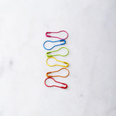 Six vibrant Cocoknits Opening Stitch Markers are arranged vertically on a light, likely marble surface. The markers in pink, blue, green, yellow, orange, and red create a visually appealing and organized display reminiscent of delicate paper clips.