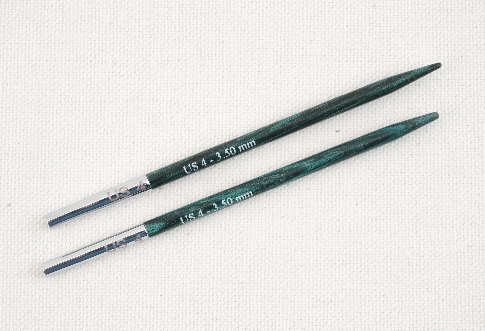 Two green-tinted wooden Dreamz Interchangeable Knitting Needle Tips by Accessories Unlimited feature smooth joins with metal connectors and are labeled "US 4 - 3.50 mm" in white text on their surface. The needles, perfect for knitting hats, are placed parallel to each other against a light beige, textured fabric background.