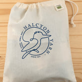 An Organic Cotton Drawstring Project Bag sits on a wooden surface. The bag proudly displays the Halcyon Yarn logo featuring a blue bird and the text "Halcyon Yarn - Yarn Fiber Tools - Since 1971." A small tag in the top right corner is marked "Eco," making it perfect as a reusable produce bag.