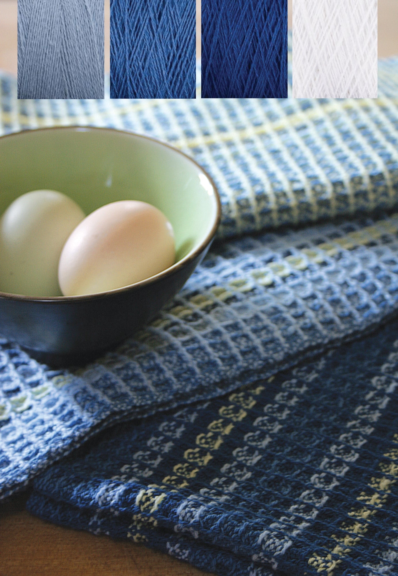 A green bowl with two eggs sits atop a table covered with woven blue and white textiles from the Halcyon Yarn Waffle Weave Dish Towel Kit. Above, close-up images showcase four yarn samples in gray, light blue, dark blue, and white, highlighting the variety available in the weaving kit.