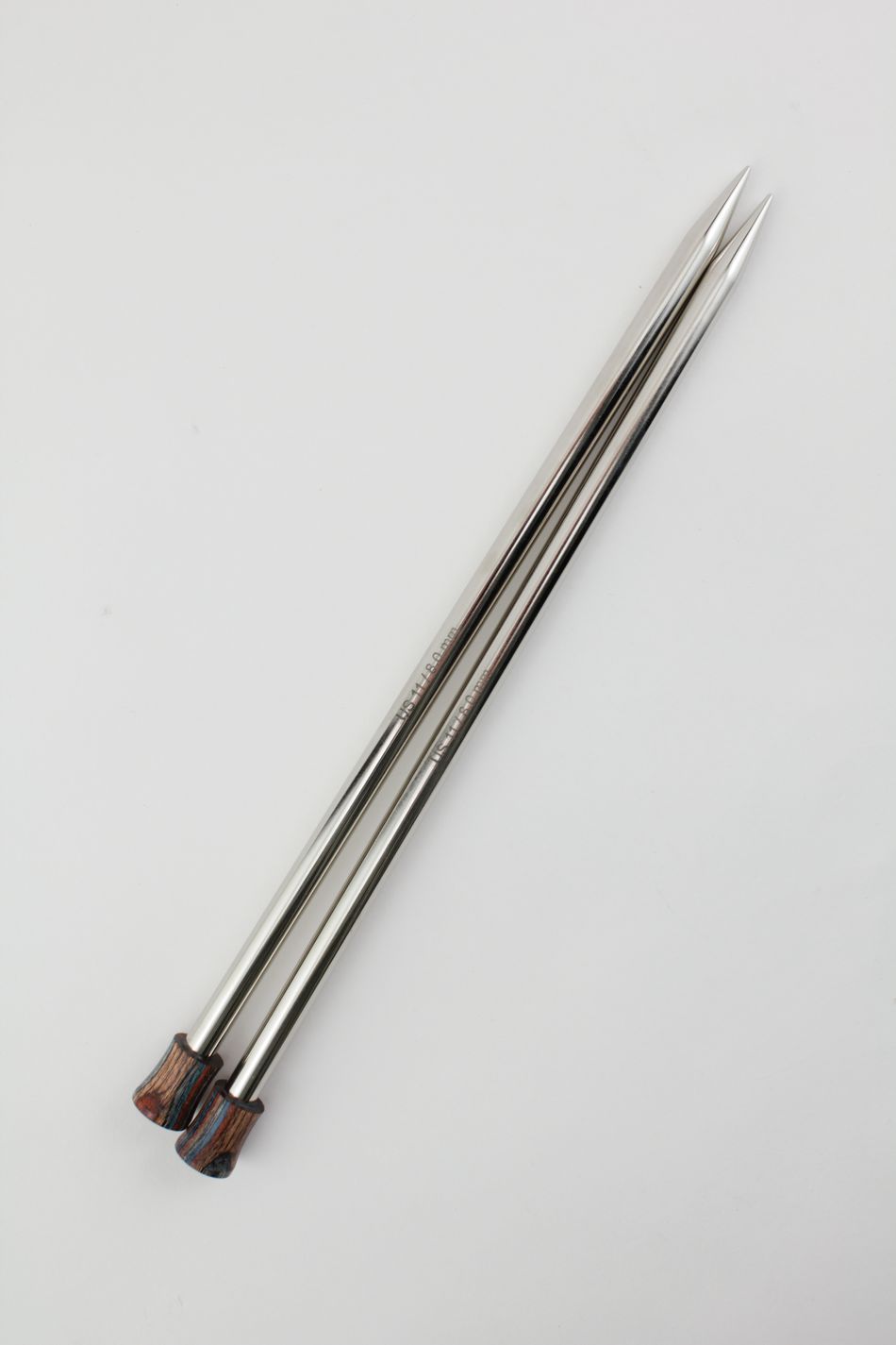 Two Nova Platina Single Point Knitting Needles from Accessories Unlimited, characterized by their long, metallic appearance resembling hollow brass pipes with pointed tips and wooden handles, lie parallel to each other on a white background.