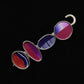 Introducing the Bubbles Shawl Pin by Bonnie Bishoff Designs: a stunning brooch featuring four oval stones set in vertical silver frames against a striking black background. Each stone is meticulously crafted with polymer clay, showcasing unique abstract patterns in shades of red, purple, and white. The top-most stone predominantly displays purple hues, while the bottom one captivates with a vibrant red and blue pattern.