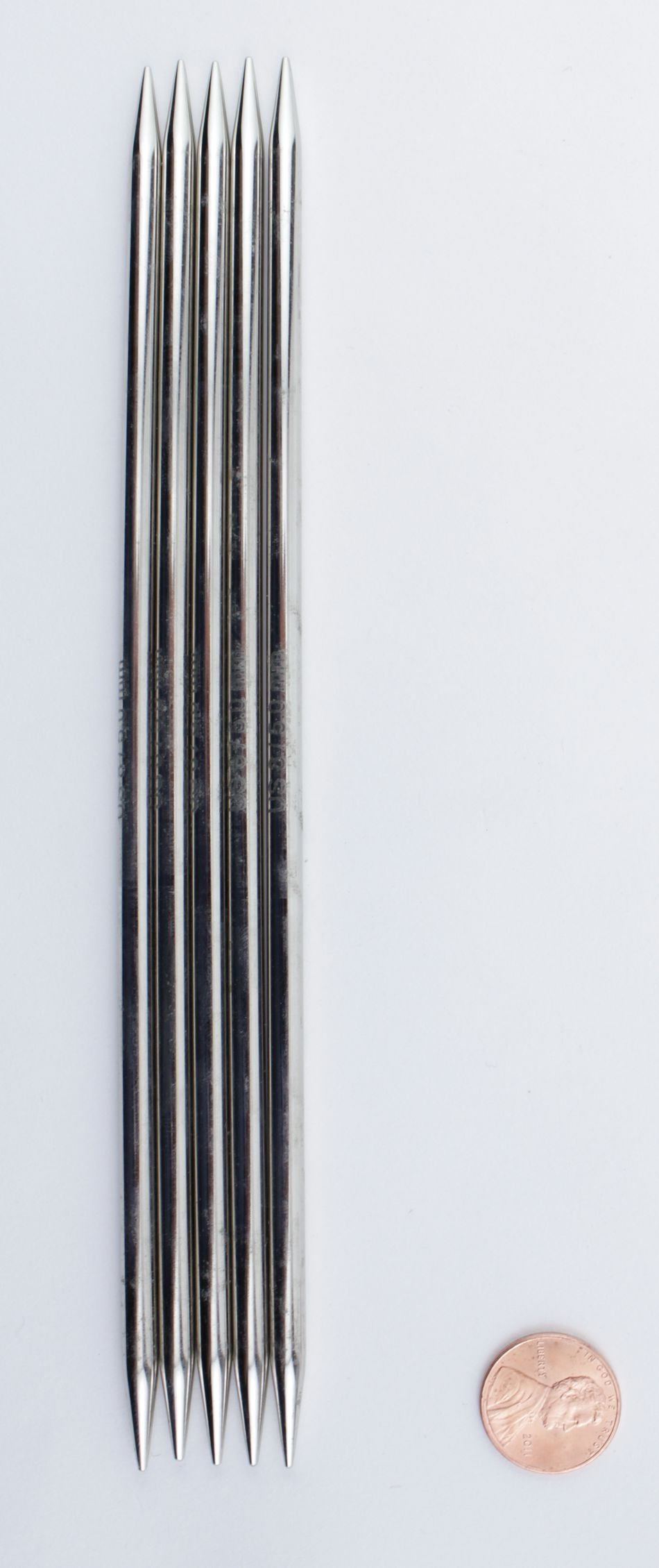 Image of six high-quality chrome, double-pointed knitting needles aligned parallel to each other next to a US penny on a white background. The needles, from the Accessories Unlimited Nova Platina Double-Point Knitting Needles range, are slightly taller than the penny, providing a size comparison.