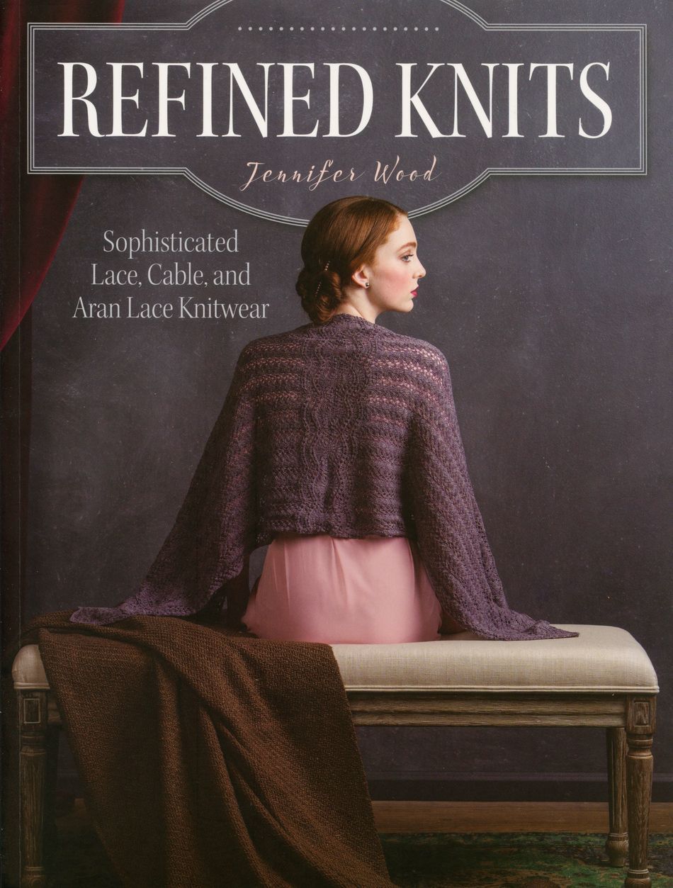 The cover of "Refined Knits" by Ingram Content, authored by Jennifer Wood, displays a seated woman adorned in a purple cable knit shawl and a pink skirt, highlighting the exquisite lace, cable, and Aran lace knitwear designs. She is seated on a bench with a folded brown knit blanket next to her.