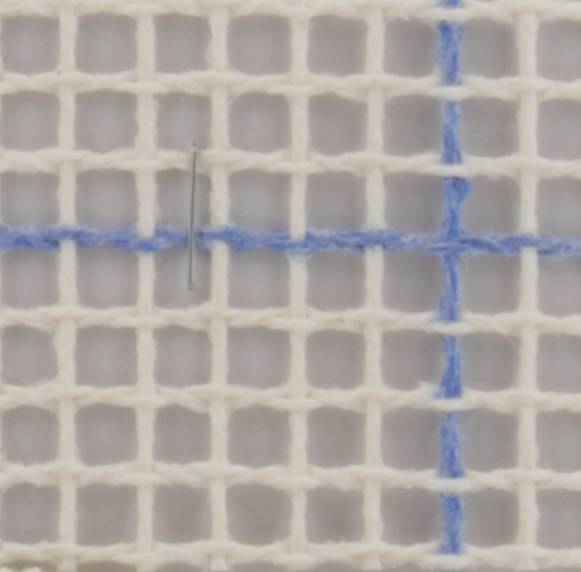 A close-up image of the Fleur de Paris Latch Hook Canvas Rug Backing, measuring 59 inches wide with a grid of 3.75 holes per inch. A silver embroidery needle is threaded through the canvas at the intersection of the blue lines, indicating it's ready for a design transfer. The canvas appears to be used for cross-stitch or needlepoint embroidery.