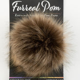 A small, fluffy ball of fur with a soft, fluffy texture in a mix of brown and black tones, attached to a black product backing card. The card reads "Furreal Pom - Extra-soft Vegan Pom Poms - Knitting Fever / Euro Yarns".