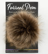 A small, fluffy ball of fur with a soft, fluffy texture in a mix of brown and black tones, attached to a black product backing card. The card reads "Furreal Pom - Extra-soft Vegan Pom Poms - Knitting Fever / Euro Yarns".