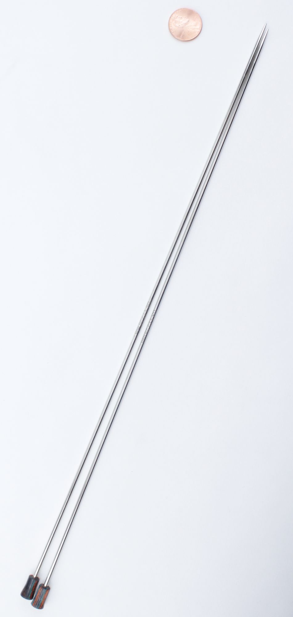 A pair of long, thin Nova Platina Single Point Knitting Needles by Accessories Unlimited are placed diagonally on a white surface. Each straight needle has a stopper on one end. A penny is positioned near the top right corner for size reference.