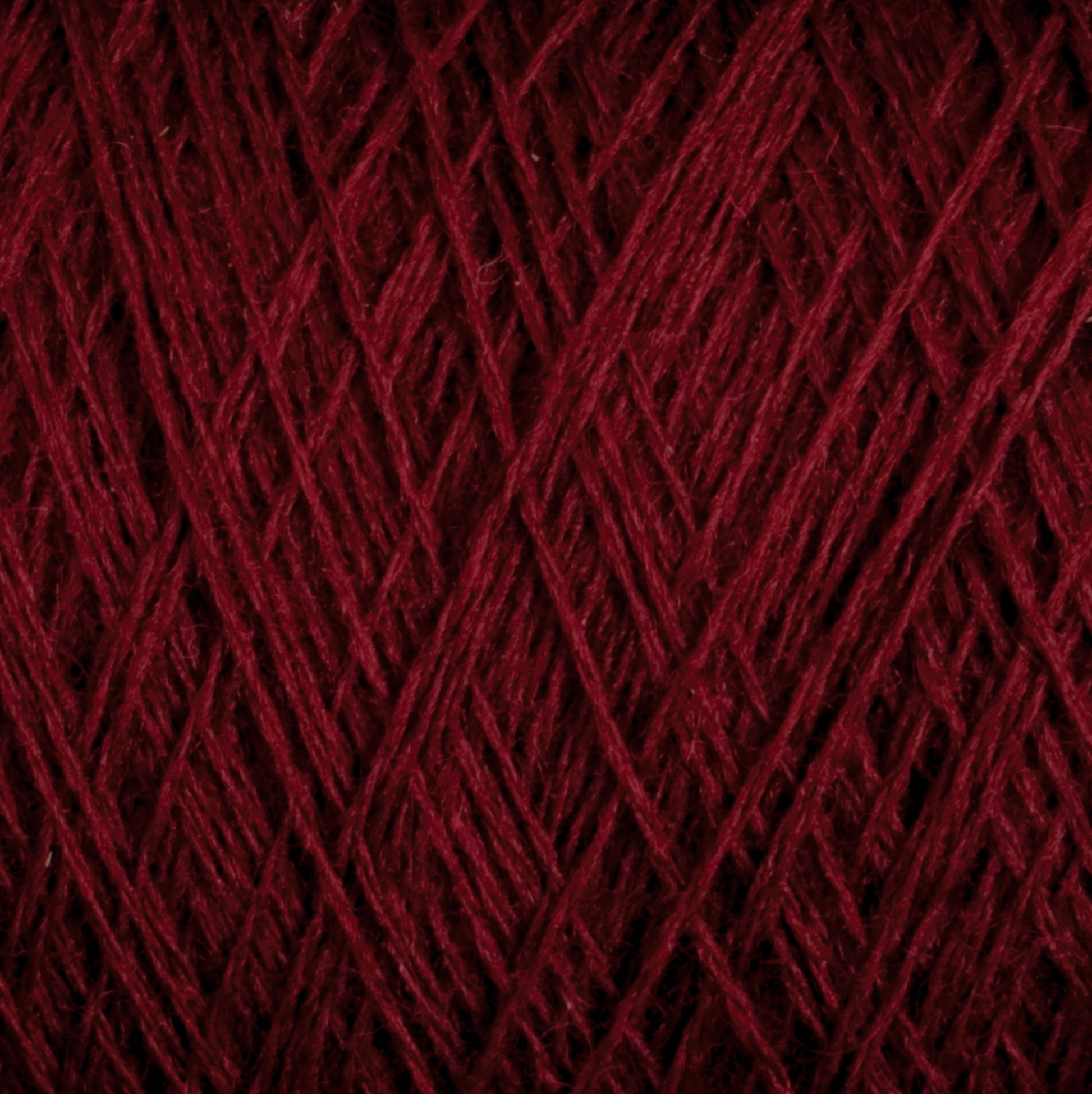 Close-up of JaggerSpun Maine Line 3/8 Yarn in maroon, manufactured by Jagger Brothers, Inc. The worsted spun yarn is tightly wound, forming an intricate pattern of overlapping strands. The texture looks soft and slightly fuzzy, with varying shades of red visible under the lighting.