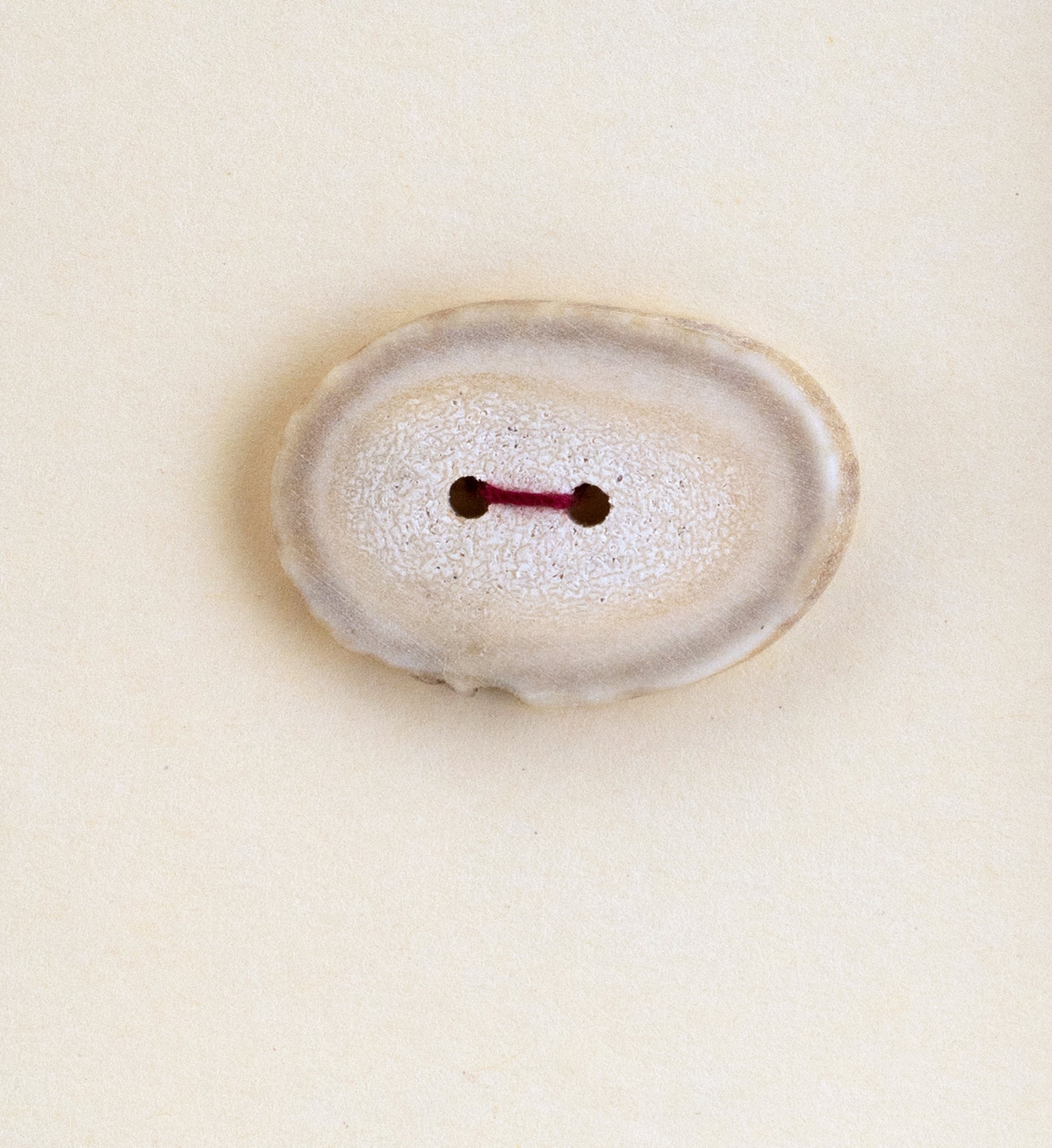 A single, oval-shaped, white button with two holes in the center, threaded with a short piece of red thread, on a plain, light-colored background. The slightly textured surface of the "Buttons - Antler" by Favour Valley Woodworking mimics the charm of wooden buttons commonly made from locally sourced materials.