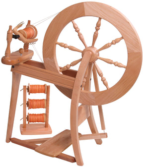 The Ashford Traditional Spinning Wheel from Ashford Handicrafts Limited features a large wheel on one side and a treadle at the base. In the foreground, there's a small stand holding three spools of orange yarn. This intricately crafted spinning wheel is polished and may include a double drive mechanism.