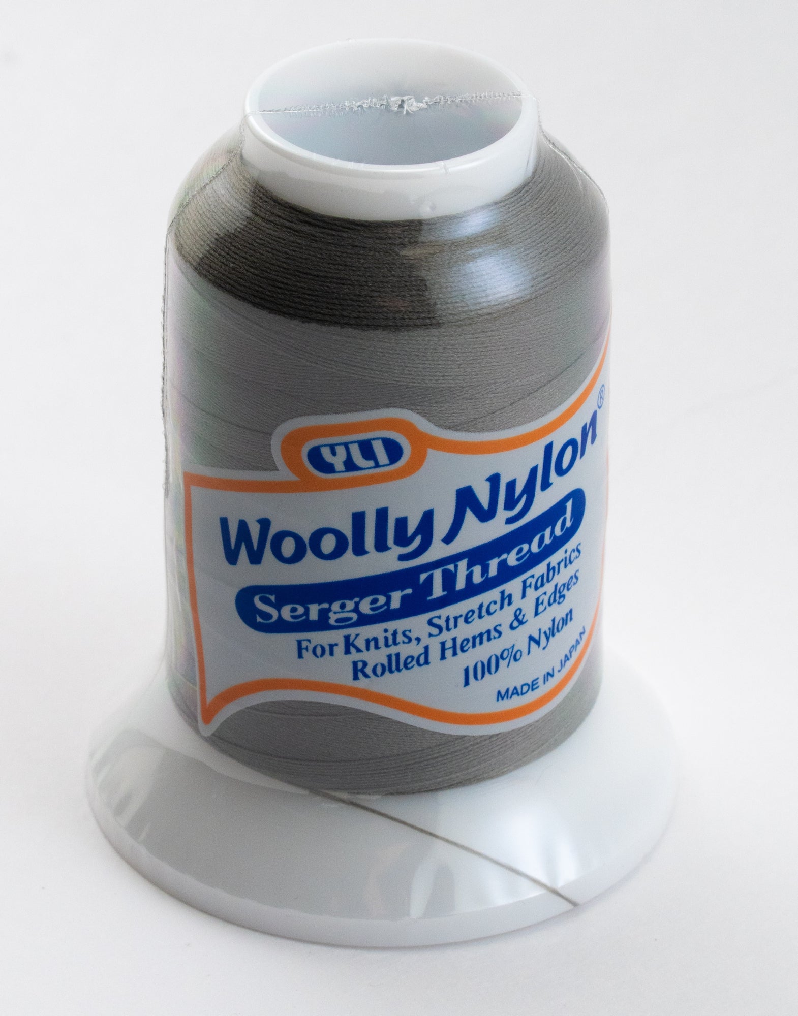 Introducing the Woolly Nylon Reinforcement Yarn by YLI Corp., a spool of high-quality serger thread in light gray, ideal for knits, stretch fabrics, rolled hems, edges, and even socks. Made from 100% nylon for exceptional durability and secure stitching. Expertly crafted in Japan, each spool is conveniently placed on a white plastic stand.