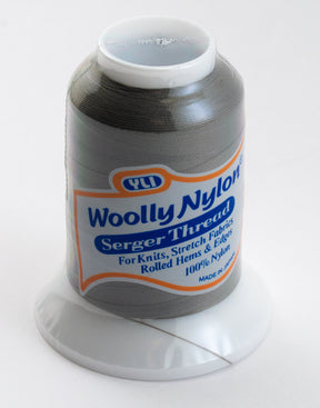 Introducing the Woolly Nylon Reinforcement Yarn by YLI Corp., a spool of high-quality serger thread in light gray, ideal for knits, stretch fabrics, rolled hems, edges, and even socks. Made from 100% nylon for exceptional durability and secure stitching. Expertly crafted in Japan, each spool is conveniently placed on a white plastic stand.