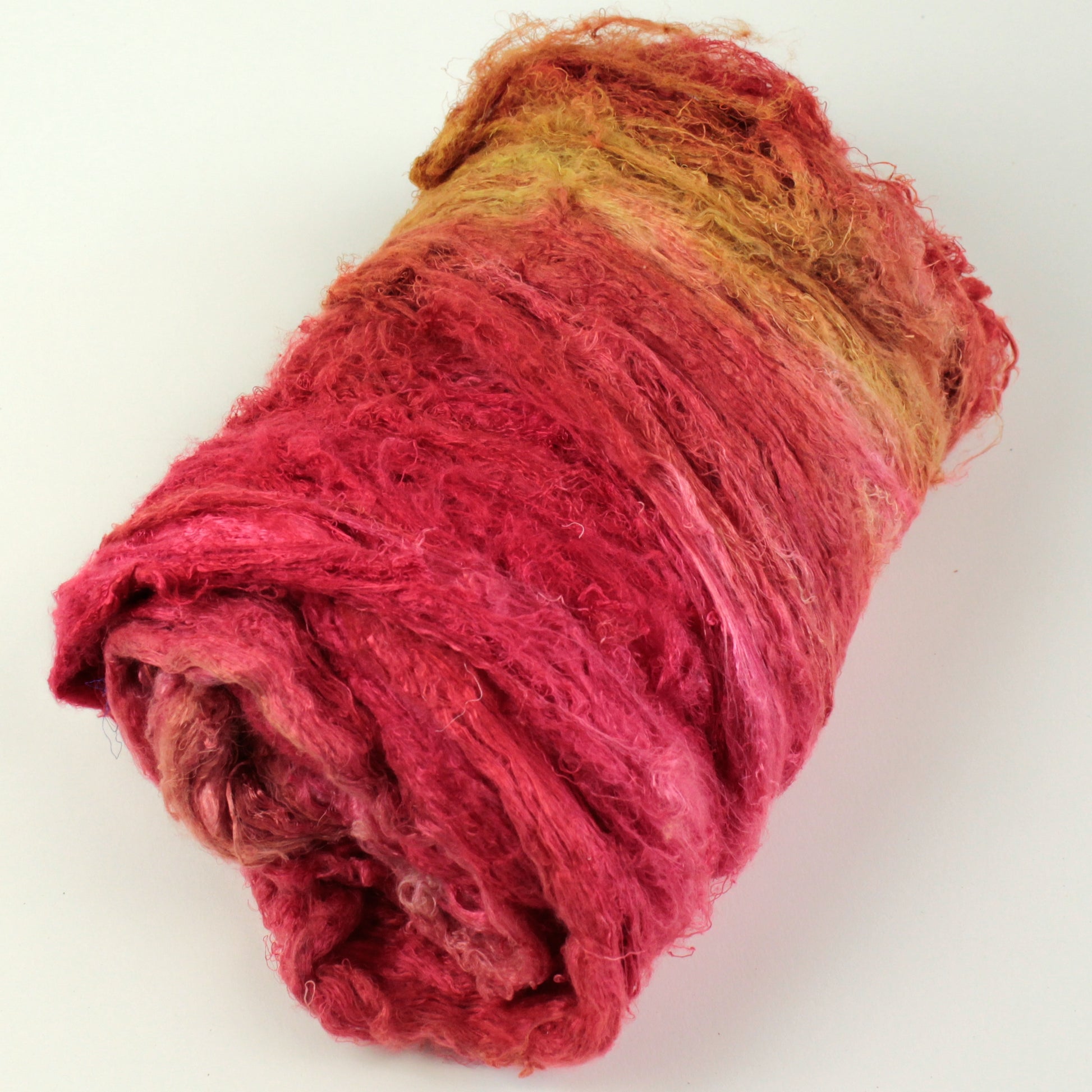 A rolled-up bundle of Demi-Carded Silk Bundles from Indian Silk Shop on a white background. The fibers appear soft and textured, with a gradient of colors blending from vibrant red to golden yellow.