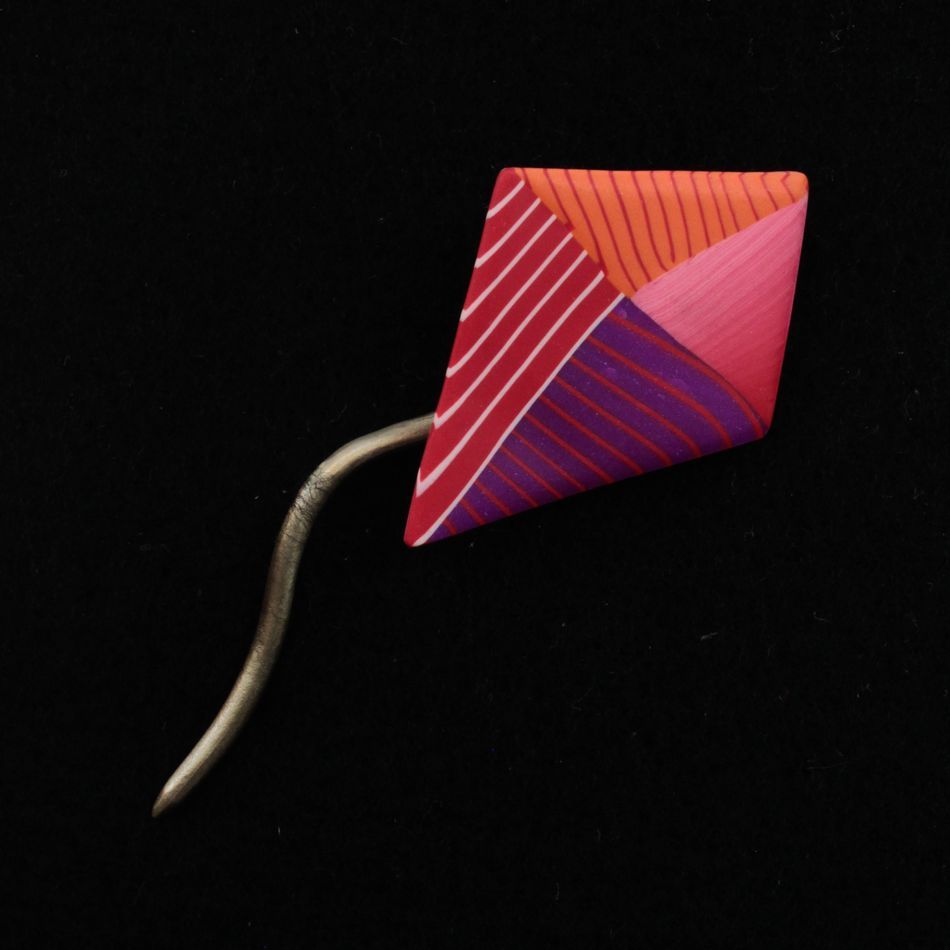 The Short Stick Shawl Pin by Bonnie Bishoff Designs is a vibrant, diamond-shaped accessory featuring a purple, pink, and red geometric pattern made from polymer clay. It has a straightforward metal fastener on the back and is perfect for use as a unique lightweight shawl closure, set against a solid black background.