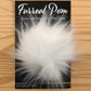 A fluffy, white faux fur pom-pom is displayed on a wooden surface. It is attached to packaging labeled "Furreal Pom by KFI," noted as "Extra-soft Vegan-fur Pom Poms." The brand "Knitting Fever / Euro Yarns" is indicated at the bottom.