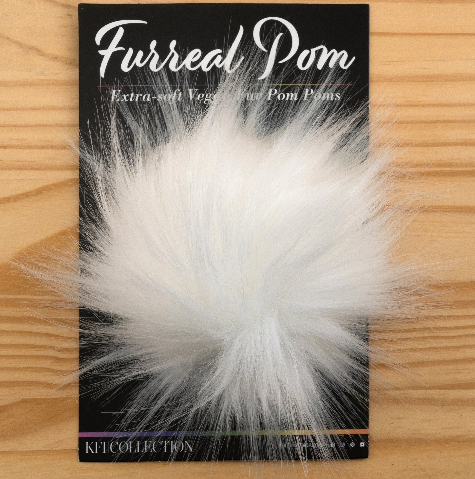 A fluffy, white faux fur pom-pom is displayed on a wooden surface. It is attached to packaging labeled "Furreal Pom by KFI," noted as "Extra-soft Vegan-fur Pom Poms." The brand "Knitting Fever / Euro Yarns" is indicated at the bottom.