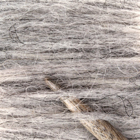 Close-up image of a tightly wrapped skein of light gray, slightly fluffy Plötulopi Unspun Icelandic Wool Yarn by Berroco, Inc., with a wooden knitting needle inserted horizontally through the strands. The yarn features subtle variations in color and visible individual fibers, giving it a soft texture.