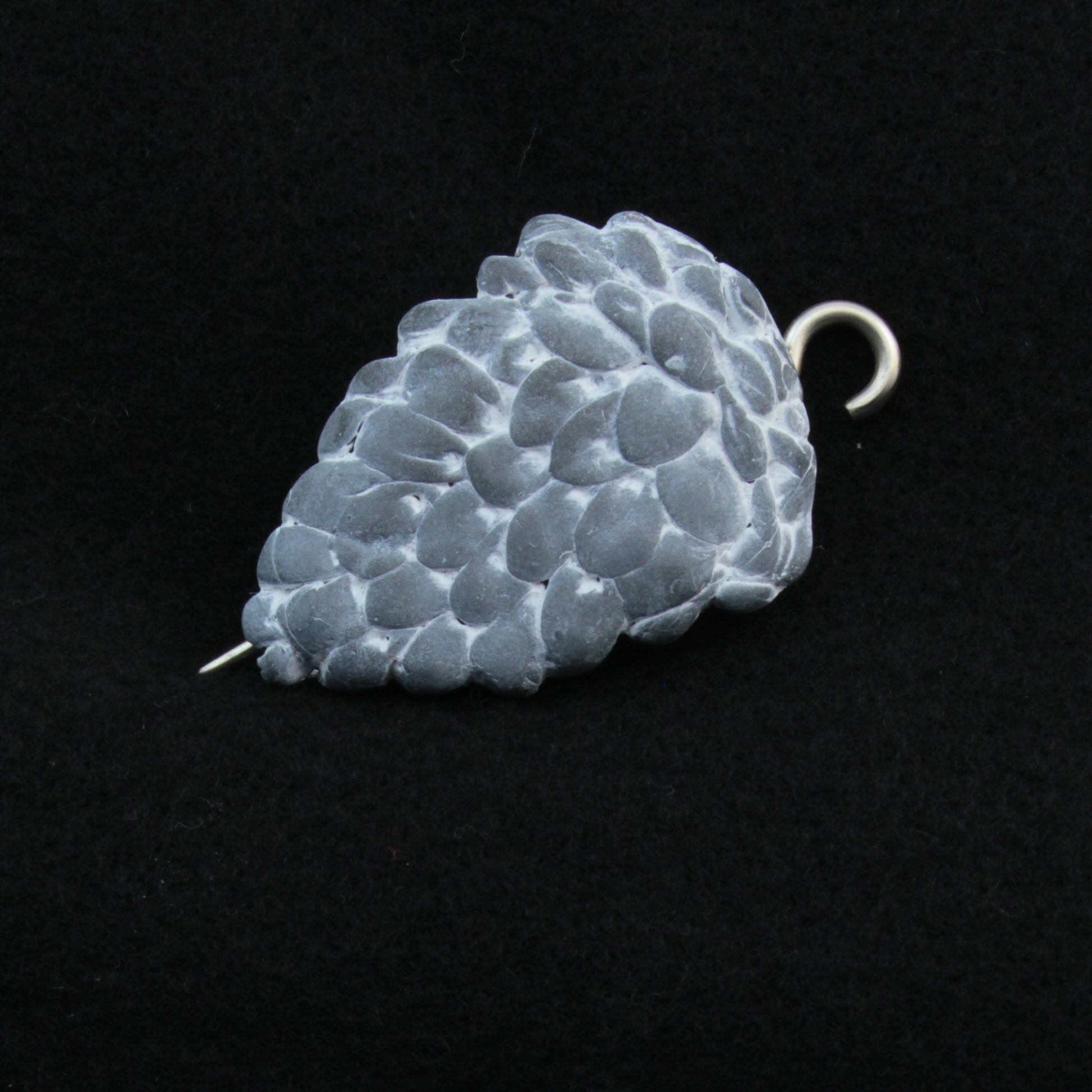 The Woodland Shawl Pin by Bonnie Bishoff Designs features a small, textured, metallic pine cone-shaped pendant with a hook at the top and a pin extending from the bottom. Crafted from lightweight polymer clay, this piece functions perfectly as an elegant shawl closure. Bonnie Bishoff ensures the pendant remains the focal point with its black background detailing.