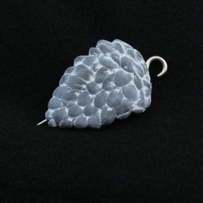 The Woodland Shawl Pin by Bonnie Bishoff Designs features a small, textured, metallic pine cone-shaped pendant with a hook at the top and a pin extending from the bottom. Crafted from lightweight polymer clay, this piece functions perfectly as an elegant shawl closure. Bonnie Bishoff ensures the pendant remains the focal point with its black background detailing.