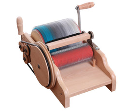 The Ashford Drum Carder - Fine 72 pts by Ashford Handicrafts Limited is a wooden carding device featuring metal carding cloth on both the large and small drums, along with a wooden handle for manual operation. It is currently being used to process fibers in red, blue, and grey through the carding drums.