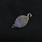 The Leaf Shawl Pin by Bonnie Bishoff Designs, crafted with intricate polymer clay design and featuring a purple and green zigzag pattern, lies against a black background. With a metal loop on each end, this elegant piece from Bonnie Bishoff is potentially perfect for attaching to jewelry or even as shawl closures.