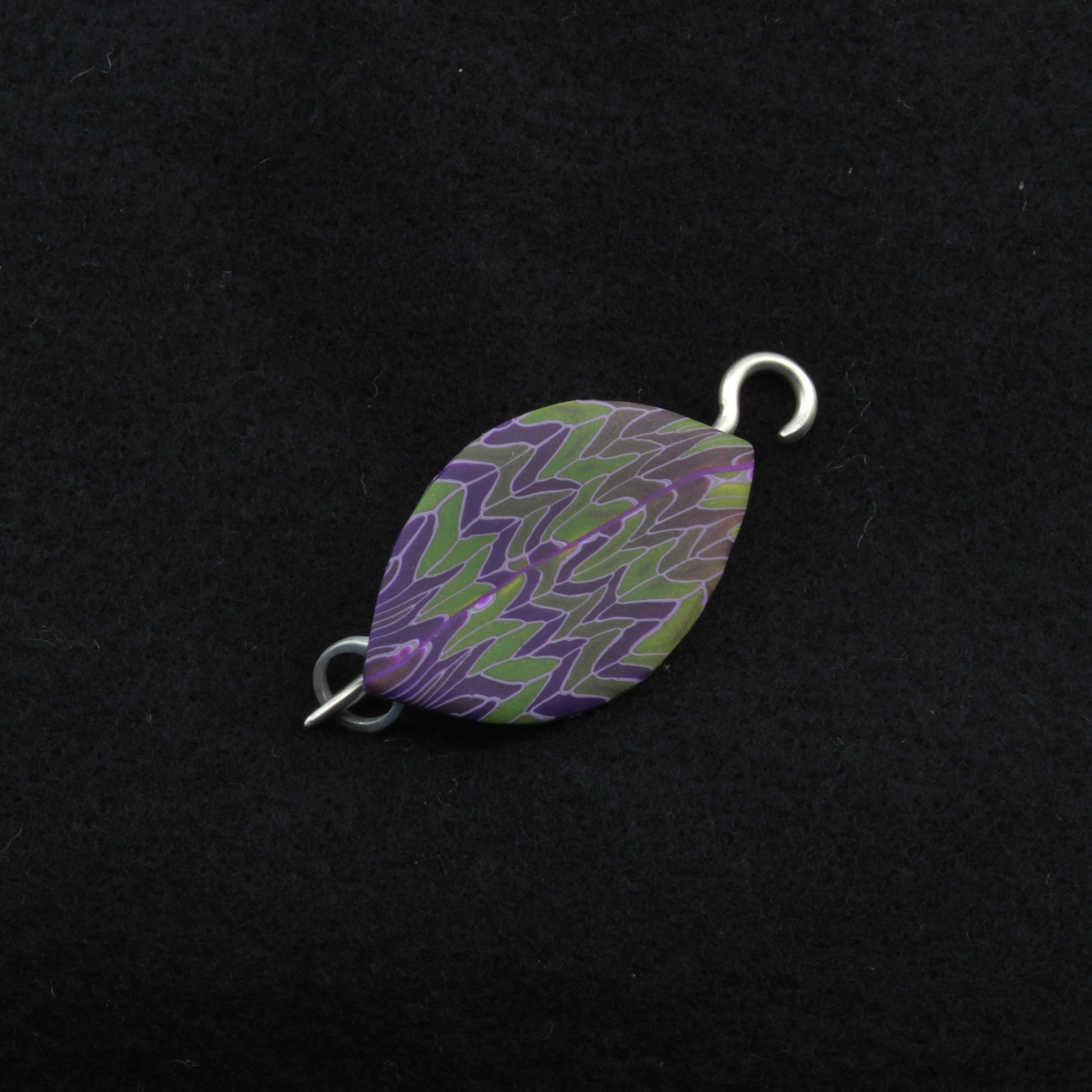 The Leaf Shawl Pin by Bonnie Bishoff Designs, crafted with intricate polymer clay design and featuring a purple and green zigzag pattern, lies against a black background. With a metal loop on each end, this elegant piece from Bonnie Bishoff is potentially perfect for attaching to jewelry or even as shawl closures.