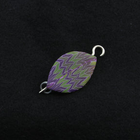 The Leaf Shawl Pin by Bonnie Bishoff Designs, crafted with intricate polymer clay design and featuring a purple and green zigzag pattern, lies against a black background. With a metal loop on each end, this elegant piece from Bonnie Bishoff is potentially perfect for attaching to jewelry or even as shawl closures.