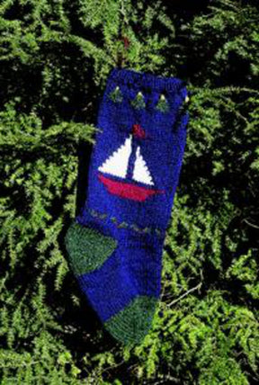 A Maine Christmas Stocking Kit by Bully Woolies, crafted from high-quality Maine spun wool, hangs gracefully from a branch of an evergreen tree. This navy blue stocking showcases a charming knitted design with a white sailboat and red hull and sail set against a blue background, accentuated by green detailing at the top and toe.