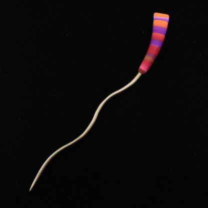 The Striped Color Stick Shawl Pin by Bonnie Bishoff Designs features a handmade, colorful layered handle crafted from polymer clay in vibrant shades of red, orange, purple, and pink. Created by Bonnie Bishoff, its metal stem boasts a wavy design that adds to its unique and artistic charm. The solid black background accentuates the intricate details of this striking hair stick.