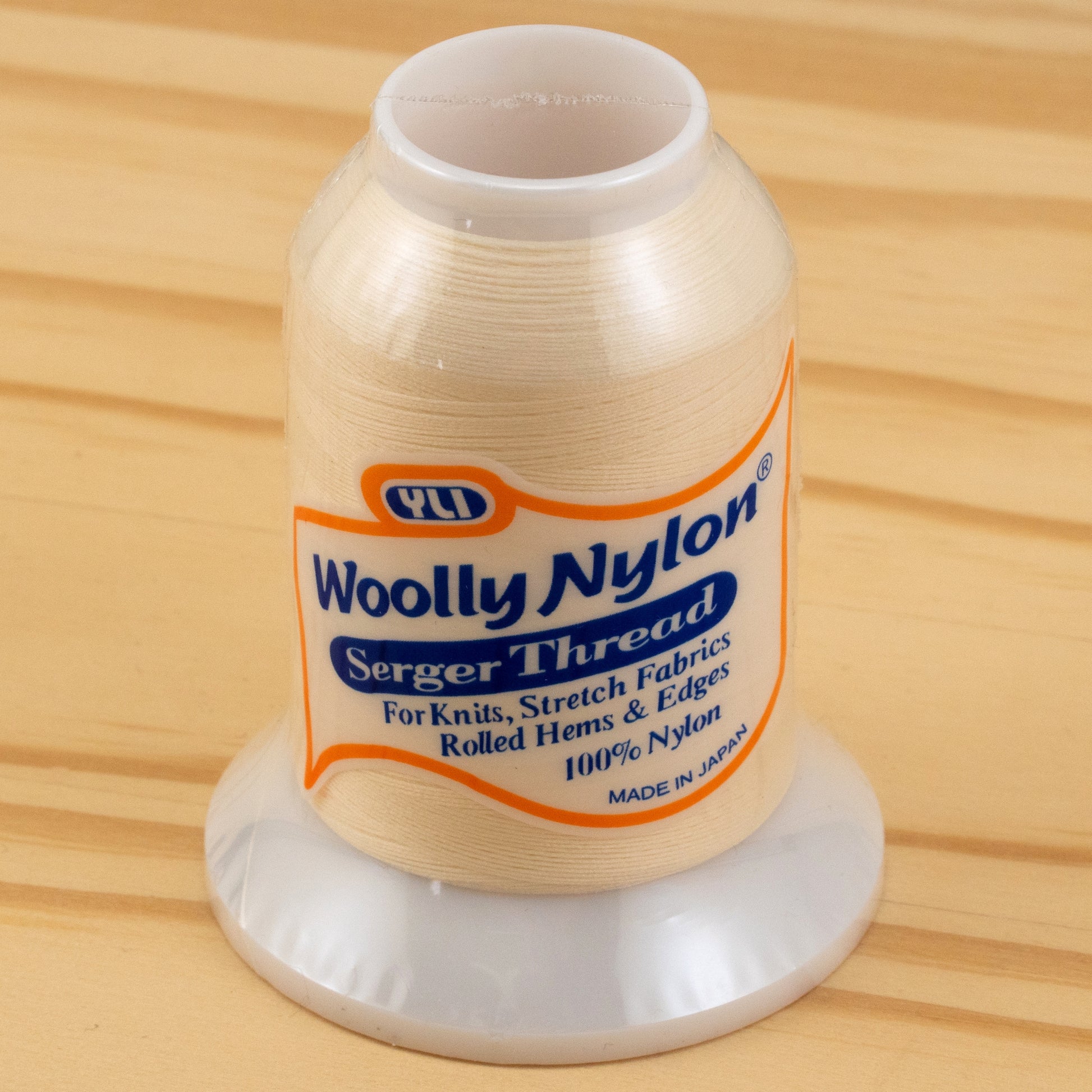 A spool of Woolly Nylon Reinforcement Yarn rests on a wooden surface, its label indicating it's perfect for knits, stretch fabrics, rolled hems, and edges. Made of 100% nylon in Japan by YLI Corp., this thread ensures the security and durability needed for crafting everything from delicate garments to sturdy socks.