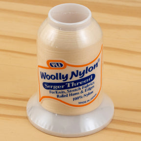 A spool of Woolly Nylon Reinforcement Yarn rests on a wooden surface, its label indicating it's perfect for knits, stretch fabrics, rolled hems, and edges. Made of 100% nylon in Japan by YLI Corp., this thread ensures the security and durability needed for crafting everything from delicate garments to sturdy socks.