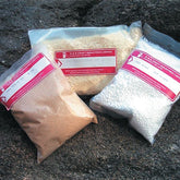 Three plastic bags containing various granular and pellet materials are placed on a rough rock surface. Each bag has a red label with white text displaying the company information. The contents, which appear to be wood shavings, powdered material, and white plastic pellets, include products such as G & K Craft's Citric Acid Crystals.