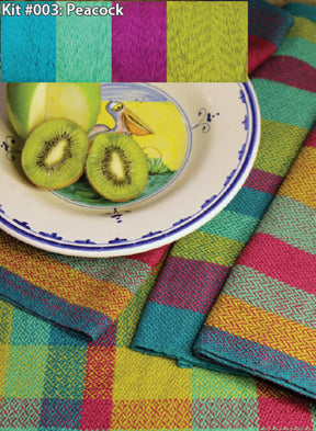 A kit labeled "Kit #003: Peacock" by Halcyon Yarn features turquoise, purple, and lime green yarns. The image showcases a ceramic plate with a pelican design, holding a sliced apple and kiwi. Two vibrant tea towels woven from organic cottolin in turquoise, magenta, and yellow are under the plate.