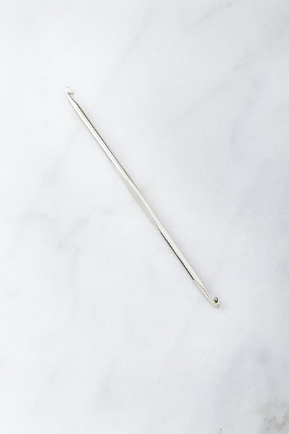 A single Cocoknits Stitch Fixer, featuring a metal crochet hook with a small hook on one end and a slight groove near the other, ideal for knitting project repairs, placed on a light, smooth surface.