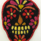 A vibrant felt sugar skull decoration set against a white background, featuring a black base adorned with bright red, yellow, and pink floral patterns, along with a yellow nose and yellow and white teeth. This intricate design captures the traditional Day of the Dead aesthetic perfectly. It would make an excellent project using The Felting Studio’s Needle Felting Tapestry Kits.