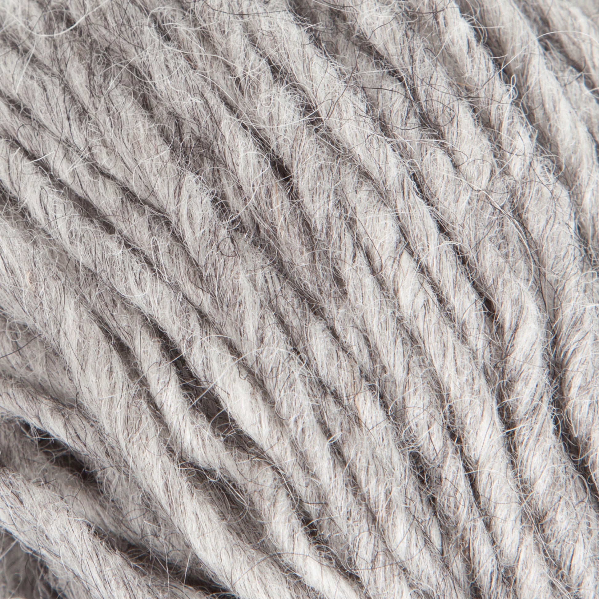 Close-up image of Jöklalopi (Bulky Lopi) yarn in grey by Berroco, Inc. showcases its soft, slightly fuzzy texture. The loosely twisted strands reveal individual fibers and their natural imperfections, exuding a rustic handmade charm. The neutral color and unique texture suggest it is ideal for knitting or crocheting cozy garments like chunky winter sweaters.