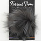 A packaging of Furreal Pom by KFI from Knitting Fever / Euro Yarns shows a fluffy, extra-soft vegan fur pom pom in a grayish-black color. The background features black cardboard with the text "Furreal Pom" and "Extra-soft Vegan Fur Pom Poms.