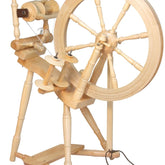 The Kromski North America Prelude Spinning Wheel is a wooden spinning wheel with a large wheel on the right side, crafted from European alder and birch. It features a foot pedal at the base and various spindles and mechanisms on the left side. This traditional single drive spinning wheel is designed for spinning fibers into thread or yarn, showcasing exceptional craftsmanship.