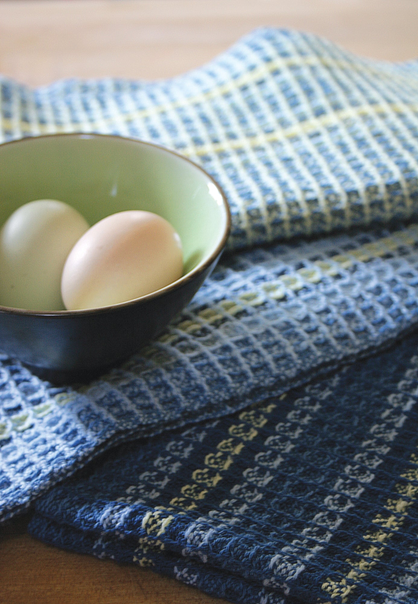 A green bowl with two eggs sits on a blue and white checkered kitchen towel from the Waffle Weave Dish Towel Kit by Halcyon Yarn, partially draped over a dark blue patterned towel from the same kit. Both towels showcase intricate waffle weave designs, resting on what seems to be a wooden surface.